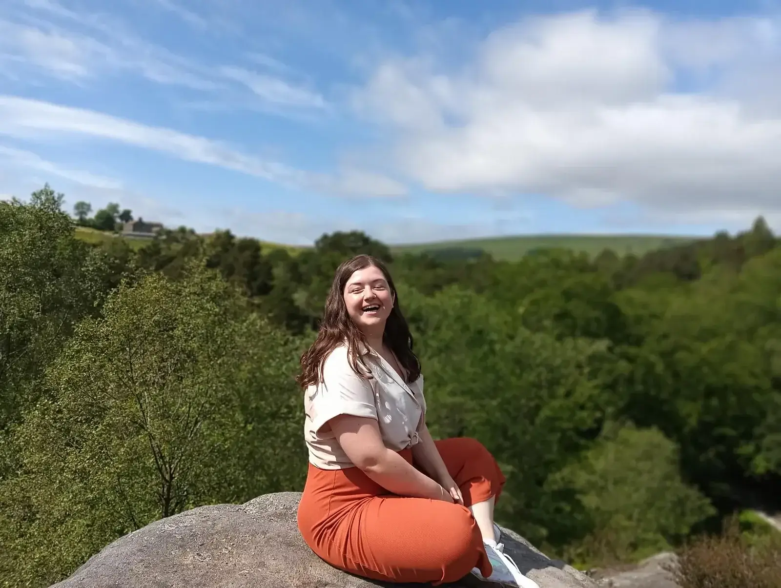 a smiling young lady sat cross legged on top of a rock with a background of trees and blue sky.