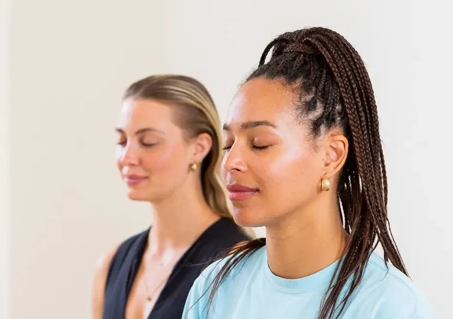 Two women, one in the foreground and one in the background, both with eyes closed, looking serene and peaceful as they are meditating.